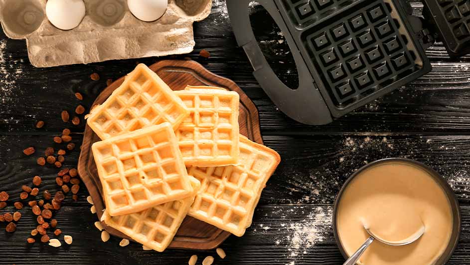 Een stapel wafels op een houten plank, met rozijnen en een kom beslag, naast een wafelijzer en eieren.