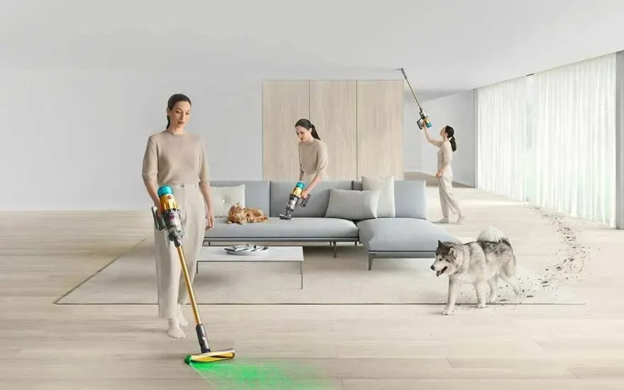 In a bright living room with large windows, three people use a Dyson cordless vacuum on the floor, sofa and ceiling, while a dog walks around.