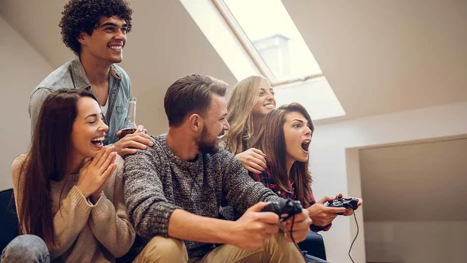 Een groep van zes vrienden speelt videogames in een kamer met een dakraam. Ze lachen en kijken geconcentreerd naar het scherm.