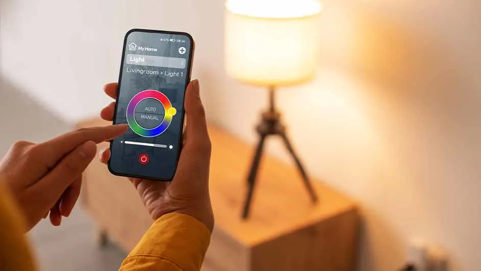 Hand bedient smartphone-app voor lampkleur, met een lamp op een houten kast op de achtergrond.