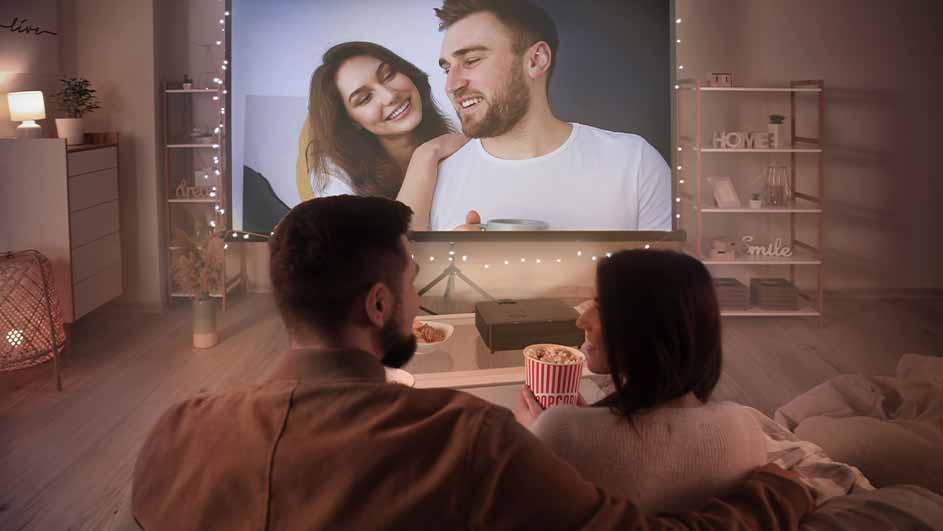 A couple watches a projector screen with a couple on it, eating popcorn in a room with shelves and soft lighting.