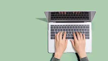 Overhead shot of hands typing on a silver laptop against a green background, with the screen reflecting keyboard.