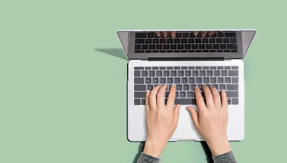 Overhead shot of hands typing on a silver laptop against a green background, with the screen reflecting keyboard.