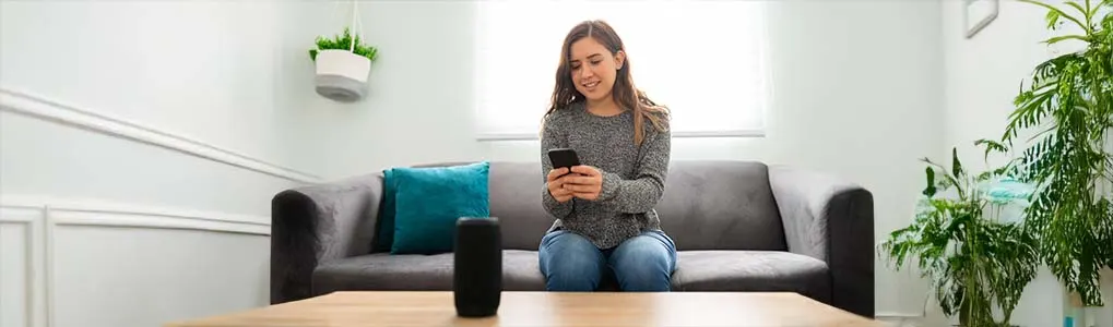 Een jonge vrouw zit op een bank en bedient een speaker met haar smartphone. De zwarte speaker staat op een tafel voor haar in een lichte woonkamer.