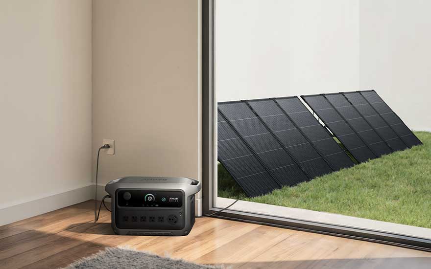 Draagbare grijze powerstation binnen op houten vloer, aangesloten op stopcontact. Buiten liggen zwarte zonnepanelen op groen gras.