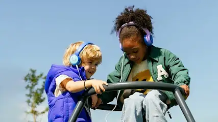 Twee kinderen met koptelefoons staan op een zwart metalen klimrek, met een blauwe lucht op de achtergrond.