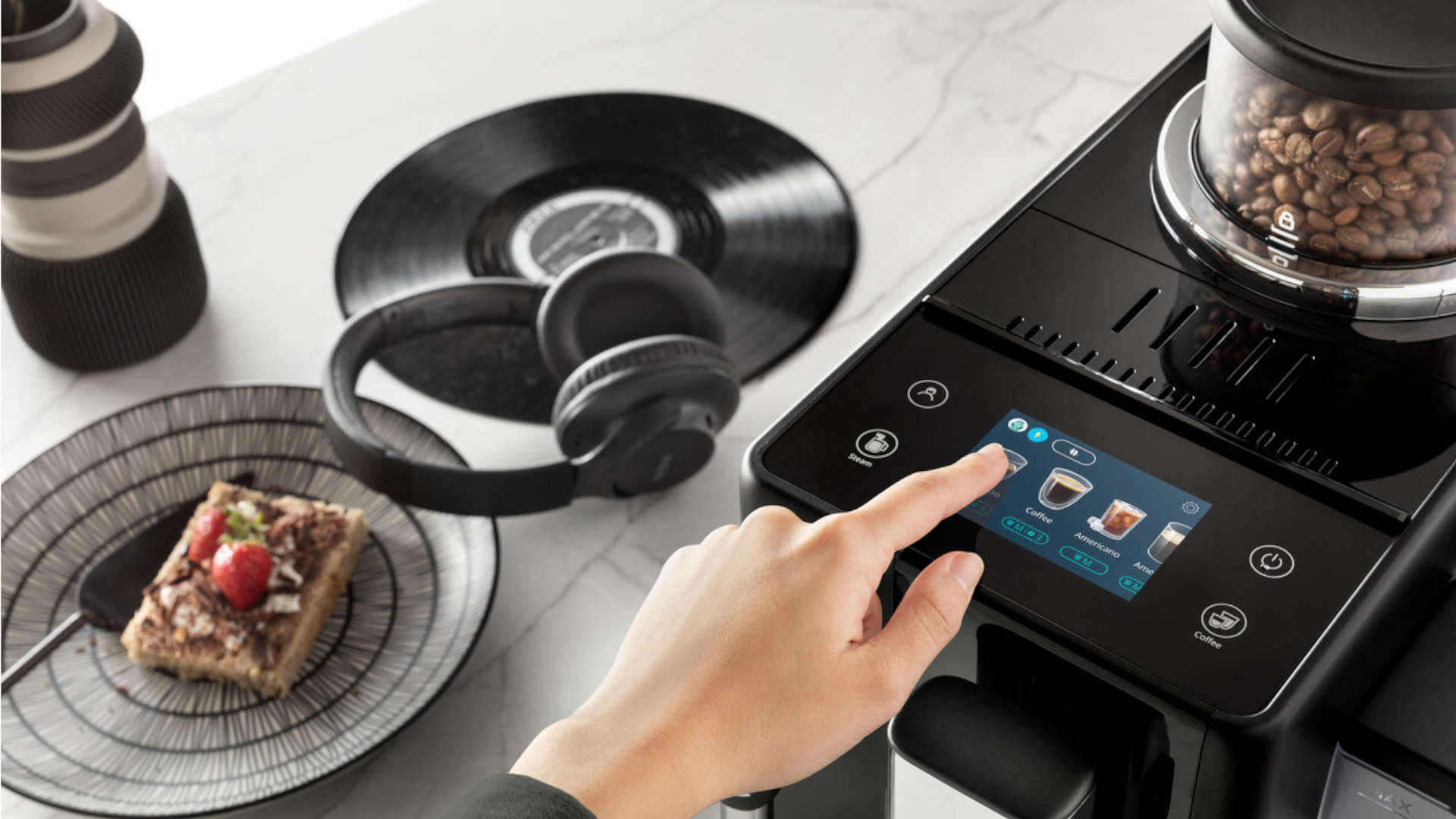 Een hand bedient een zwart koffiezetapparaat met bonenreservoir. Er staat een bord met gebak, koptelefoon en een vinylplaat.