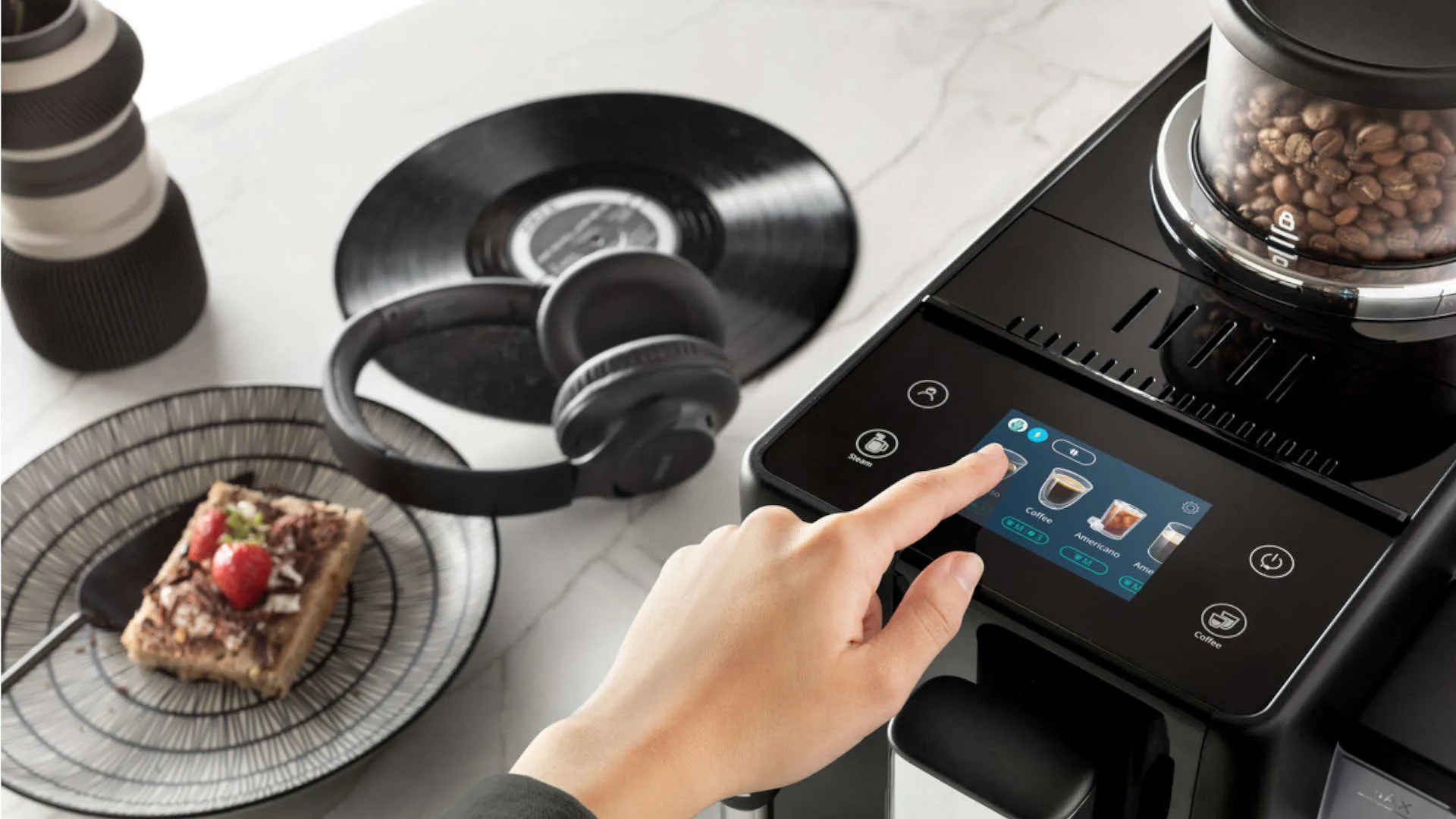 Een hand bedient een zwart koffiezetapparaat met bonenreservoir. Er staat een bord met gebak, koptelefoon en een vinylplaat.