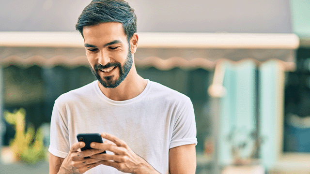 Een man in een wit shirt staat buiten en glimlacht terwijl hij naar een mobiele telefoon in zijn handen kijkt.