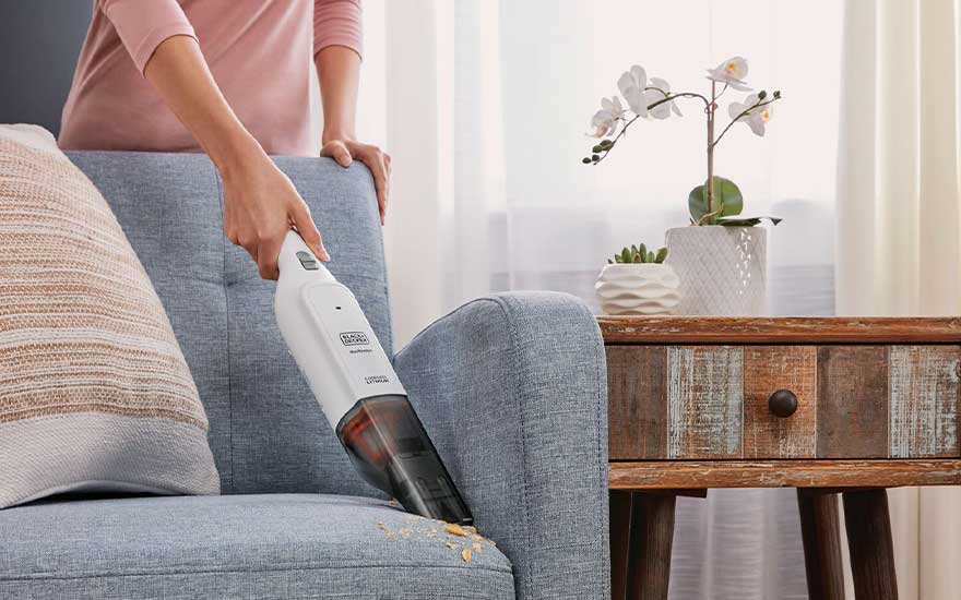 A handheld vacuum removes crumbs from a light blue sofa, beside a rustic wooden side table with cushions and a white orchid.