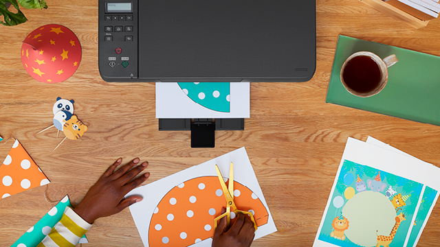 Bovenaanzicht van een houten bureau met printer, kopje, schaar, papier en decoraties voor een feestje.