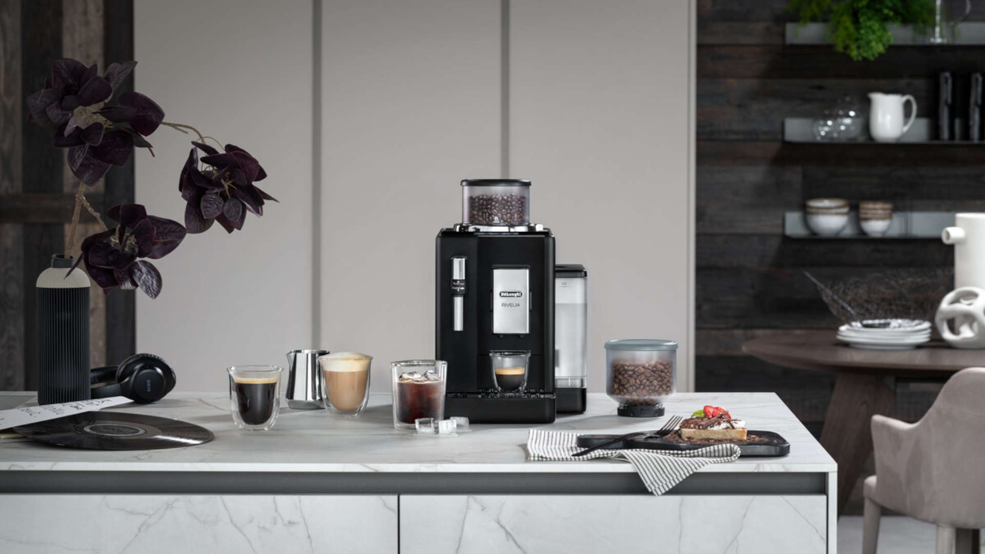 Een zwarte koffiemachine op een marmeren aanrecht met koffie, een toetje, een hoofdtelefoon en een bloemstuk.