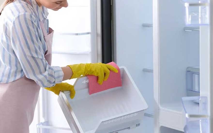 A woman wearing rubber gloves scrubs a fridge drawer with a pink sponge while the inside of the refrigerator is completely empty.