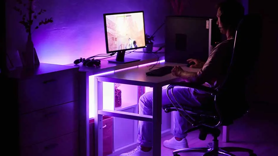 Man zit aan een bureau met een computer, toetsenbord en muis in een kamer met paarse verlichting.