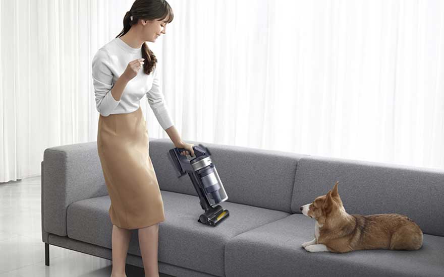 Een vrouw verwijdert dierenharen van de bank met een turboborstel, terwijl haar hond ontspannen toekijkt.