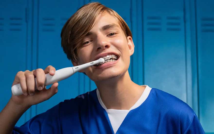 Een tiener met beugel poetst zijn tanden met een witte elektrische tandenborstel voor blauwe lockers en glimlacht zelfverzekerd.
