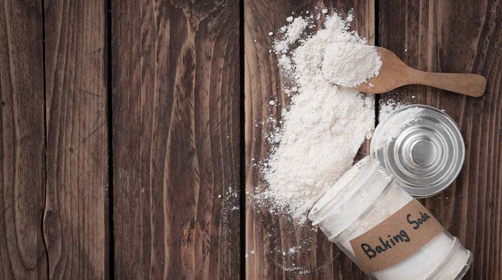 A wooden table has a spilled powder, a spoon, a metal lid, and a jar labeled "Baking Soda" on it.