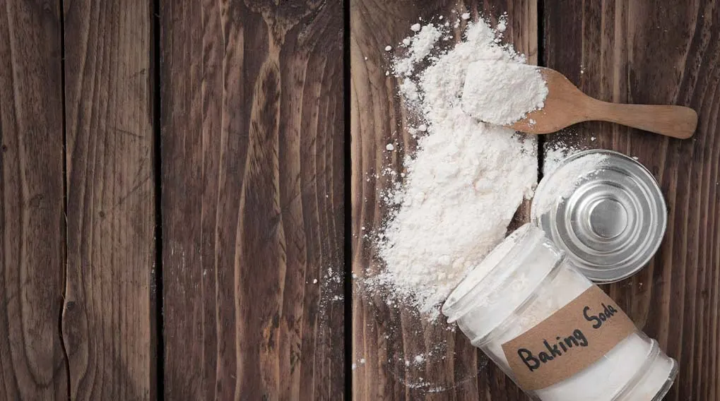 A wooden table has a spilled powder, a spoon, a metal lid, and a jar labeled "Baking Soda" on it.