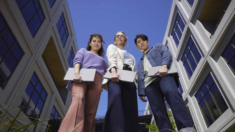 Drie mensen met laptops staan buiten voor gebouwen met blauwe ramen tegen een blauwe hemel.