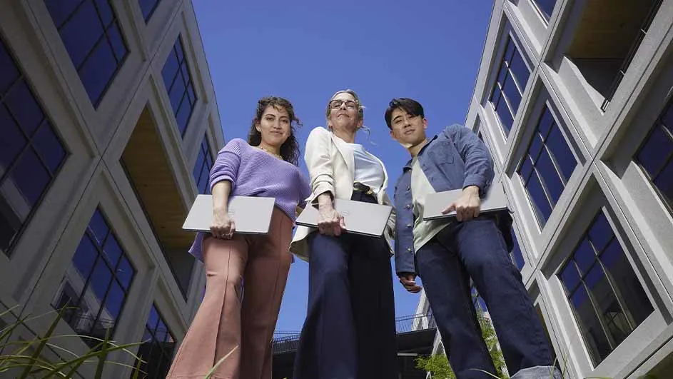 Drie mensen met laptops staan buiten voor gebouwen met blauwe ramen tegen een blauwe hemel.