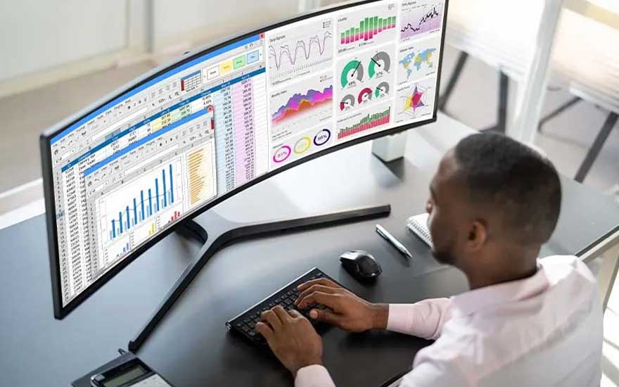 An employee analyses spreadsheets and vivid dashboards on a curved ultrawide monitor while typing on a compact wireless keyboard.