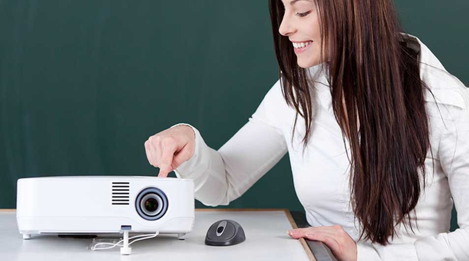 A woman with long brown hair and a white top is sitting at a table, pressing a button on a white projector. No brand name is visible on the device.