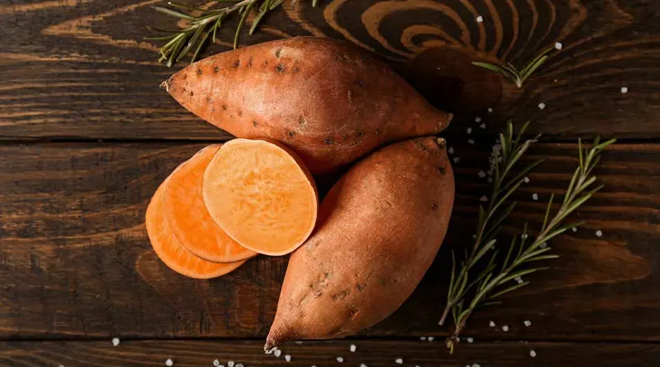 Twee hele zoete aardappelen en enkele plakjes liggen op een houten tafel met rozemarijn en zout.
