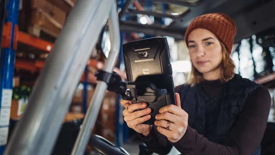 Een vrouw in een pakhuis bedient een zwarte tablet op een heftruck, met schappen op de achtergrond.