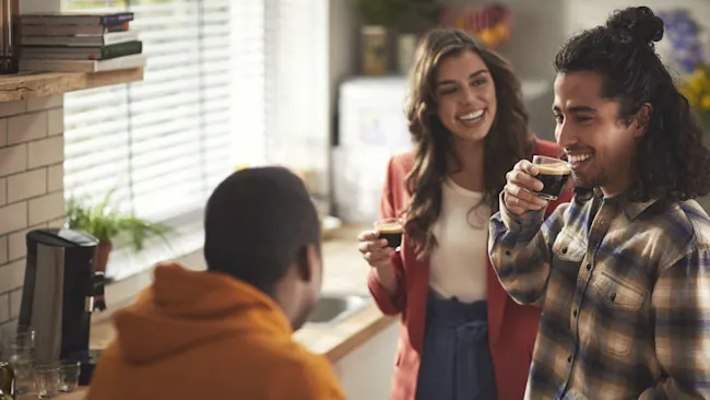 Drie mensen in een keuken. Twee glimlachen terwijl ze koffie drinken, een derde staat met de rug naar de camera.