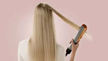 Een vrouw met lang, blond haar gebruikt een stijltang tegen een roze achtergrond.