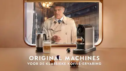 Reclamefoto toont een koffiemachine, melkopschuimer, glas koffie en capsules op een bruine tafel met een man op de achtergrond.