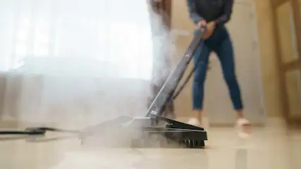 Een persoon stoomt een beige tegelvloer met een zwarte stoomreiniger. Er is witte stoom zichtbaar bij de reiniger en een raam.