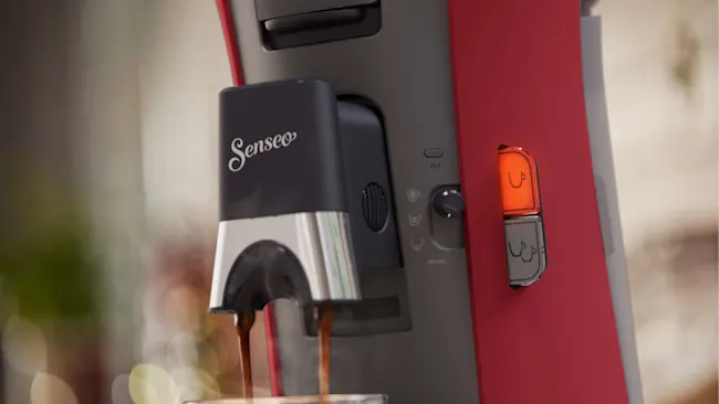 Senseo koffiezetapparaat schenkt koffie. Het apparaat is rood en grijs met knoppen voor instellingen.