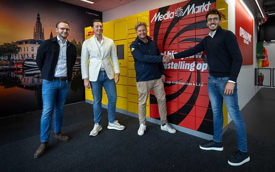 Vier mannen staan glimlachend voor DHL- en MediaMarkt-pakjeskluizen. Twee van hen schudden elkaar vriendelijk de hand.
