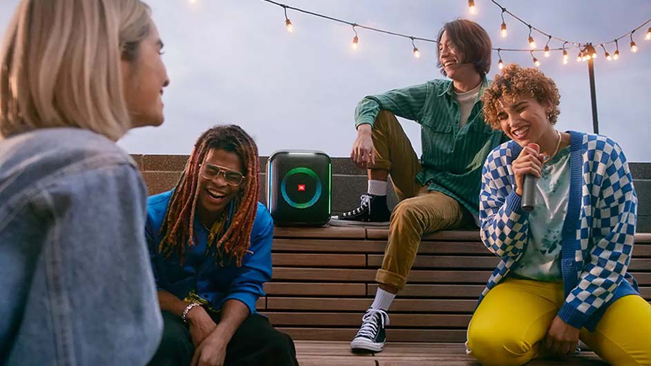 Vijf mensen zitten buiten met een microfoon en een luidspreker met lichtjes, en een lichtsnoer hangt erboven.