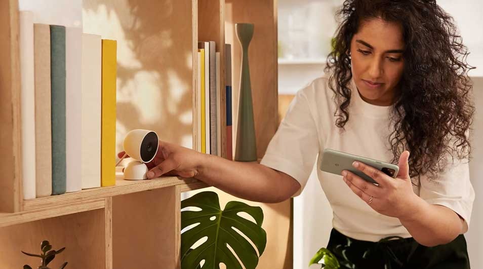 Een vrouw plaatst een witte Google Nest Cam op een plank, terwijl ze haar smartphone vasthoudt. De camera is compact en modern vormgegeven.