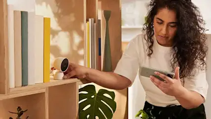 Een vrouw plaatst een witte Google Nest Cam op een plank, terwijl ze haar smartphone vasthoudt. De camera is compact en modern vormgegeven.