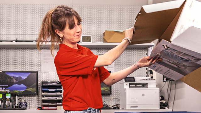 Een vrouw in een rode blouse pakt een kartonnen doos uit in een kantooromgeving met een printer en computerscherm.