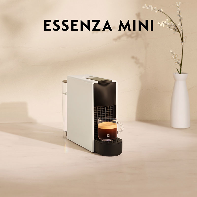 Een witte Essenza Mini koffiemachine met een kop koffie, tegen een neutrale achtergrond met bloemen in een witte vaas.