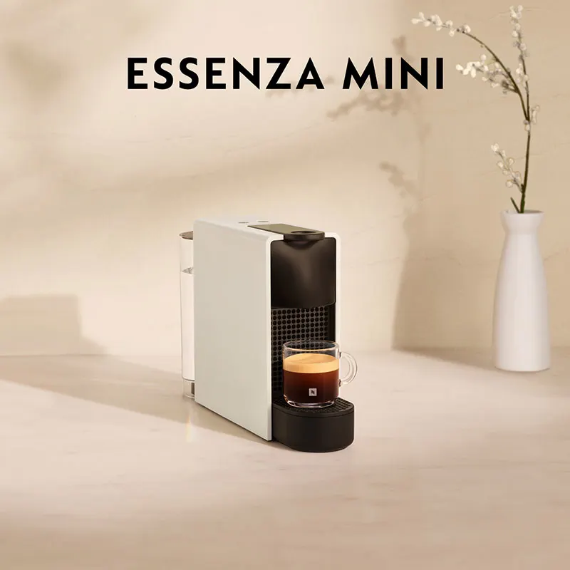 Een witte Essenza Mini koffiemachine met een kop koffie, tegen een neutrale achtergrond met bloemen in een witte vaas.