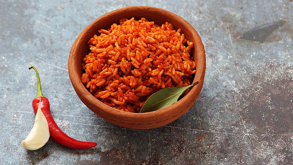 Een bruine kom gevuld met oranje rijst staat naast een rode peper en een knoflookteentje op een grijze ondergrond.