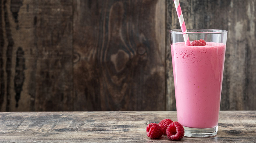 Roze smoothie in glas met rietje en framboos, op houten tafel. Drie frambozen liggen los op de voorgrond.
