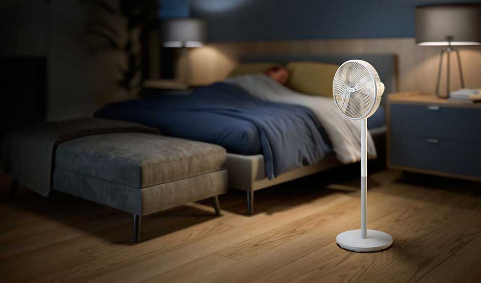 A fan stands beside a bed in a dark bedroom, providing cooling comfort during the night’s rest.