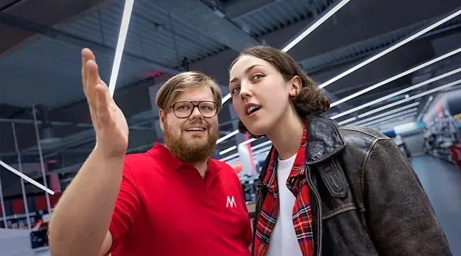 Two people stand in a store. The person on the left wears a red shirt, and glasses and raises their hand. The other is in a jacket.