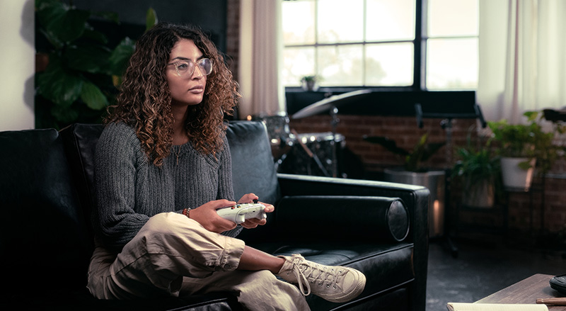 Persoon in groene trui houdt witte Xbox-controller vast. Donkere broek, sneakers en houten vloer creëren een ontspannen gaming-sfeer.