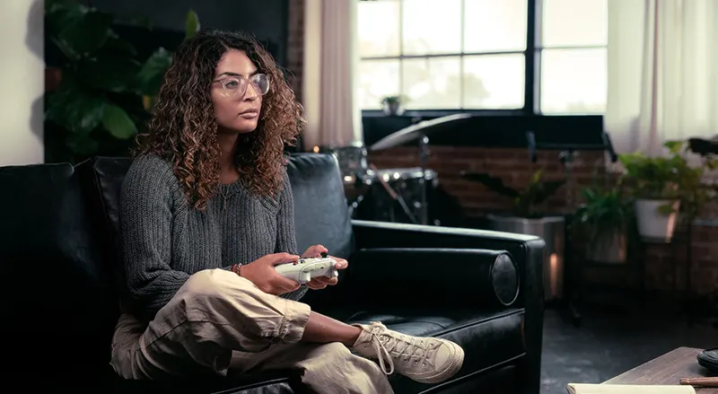 Persoon in groene trui houdt witte Xbox-controller vast. Donkere broek, sneakers en houten vloer creëren een ontspannen gaming-sfeer.