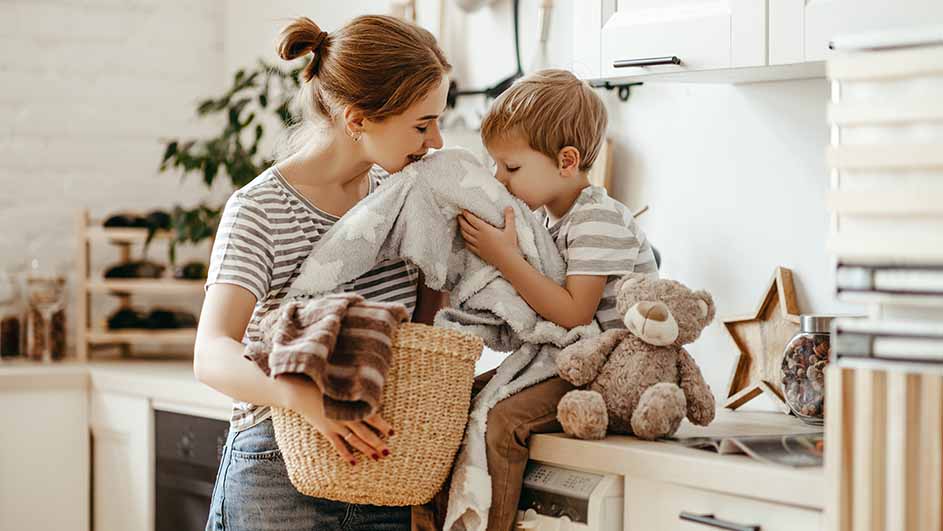 Een vrouw met een mand vol wasgoed staat naast een kind met een teddybeer in een keuken.