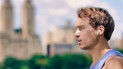 Een man met een blauw hemd en oortjes kijkt naar links, met gebouwen en groen op de achtergrond.