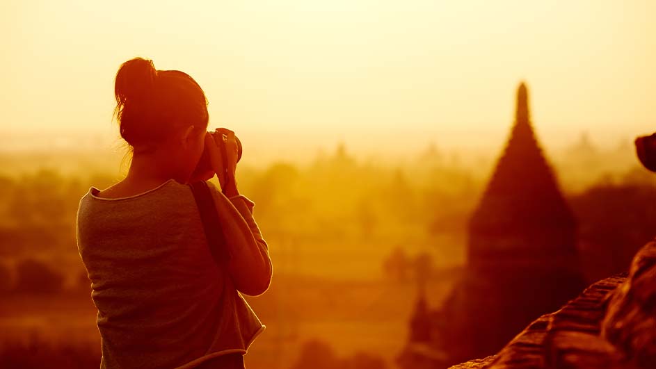 Een vrouw met een camera staat op een heuvel. Op de achtergrond zijn tempels en bomen te zien bij zonsondergang.