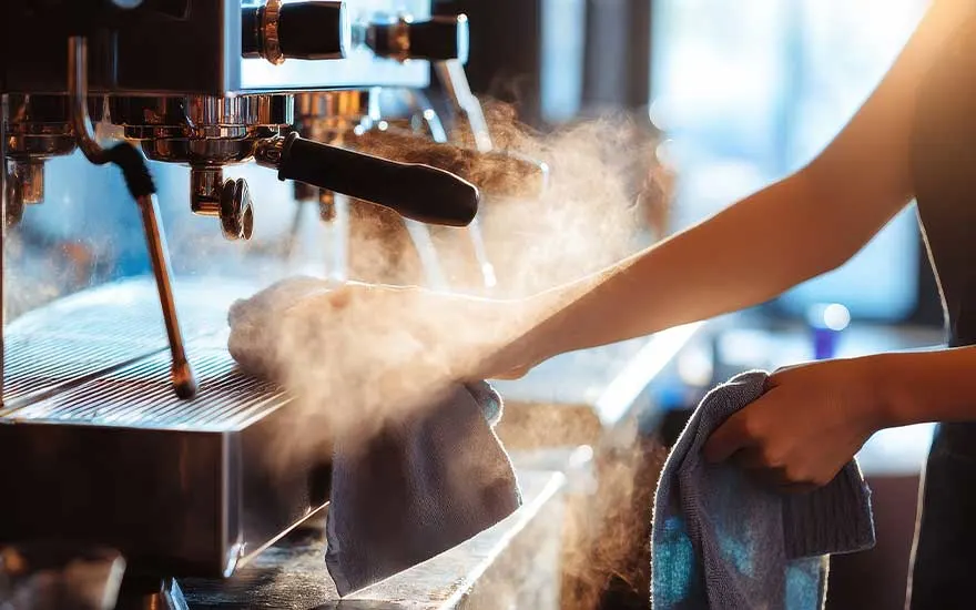 Someone cleans a hot professional espresso machine as steam swirls around the metal parts in the warm café lighting.